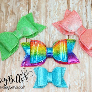 May include: Four glitter hair bows in different colors and styles. The bows are green, pink, rainbow, and blue. The bows are on a wooden background.