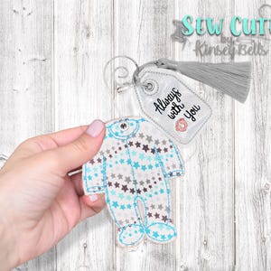 May include: A white tag with a blue and gray baby onesie design and the text "Always with you". The tag is attached to a gray tassel.