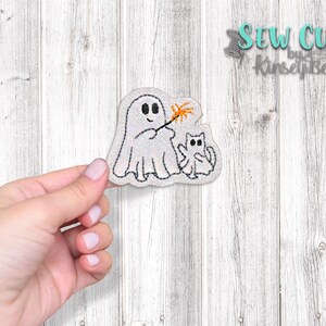 May include: Embroidered patch featuring a white ghost holding a sparkler and a small white cat. The patch is on a white wood background.