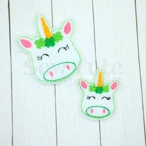 May include: Two white felt unicorn head appliques with green shamrock crowns and orange horns. The larger applique has a pink nose and pink eyes. The smaller applique has a pink nose and black eyes.