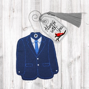 Op de afbeelding: Een marineblauw, pakvormig ornament met een witte kraag en blauwe stropdas. Een label met de tekst "Always with You" en een rode vogelillustratie hangt naast het pak. Een grijze kwast is aan het label bevestigd.