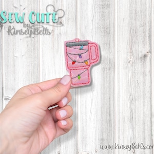 May include: Pink embroidered mug with a string of colorful lights hanging from the handle. The mug is on a white wooden background.