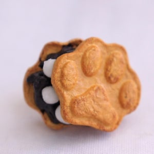 以下が含まれることがあります： 明るい茶色の外観と白いドットのある暗いフィリングの肉球型クッキー。クッキーは肉球のデザインで、3つの明確な肉球パッドの印象があります。クッキーはおやつです。