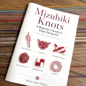 May include: A white instructional book titled Mizuhiki Knots: A Beginner's Guide to Paper String Art, featuring illustrations of six knot styles including Ume Blossom and Cobblestone, with colorful paper strings arranged horizontally in the background.
