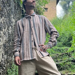 May include: A person wearing a striped, light brown and blue shirt, beige pants, and a gray beanie stands on stone steps in a lush green forest setting.