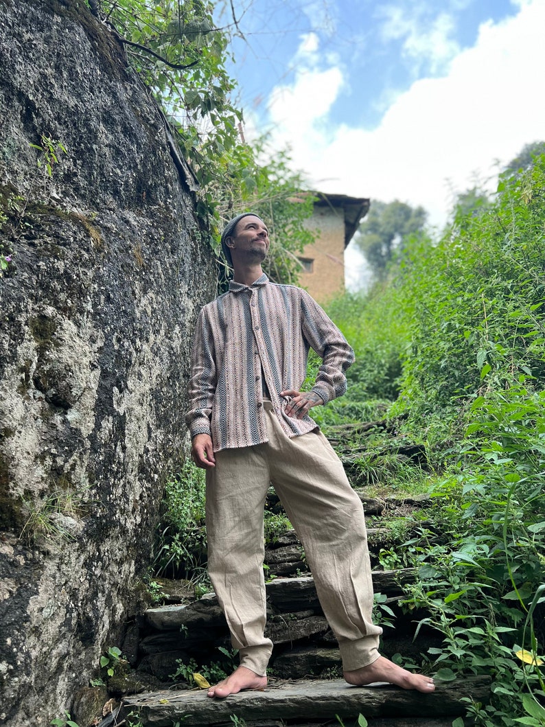 May include: A person wearing a striped, light brown and blue shirt, beige pants, and a gray beanie stands on stone steps in a lush green forest setting.