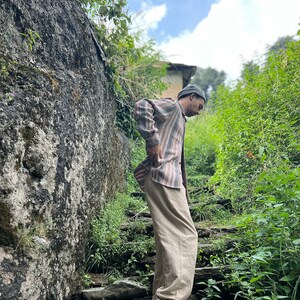May include: A person wearing a striped shirt and beige pants stands on a set of stone steps in a lush green forest setting.