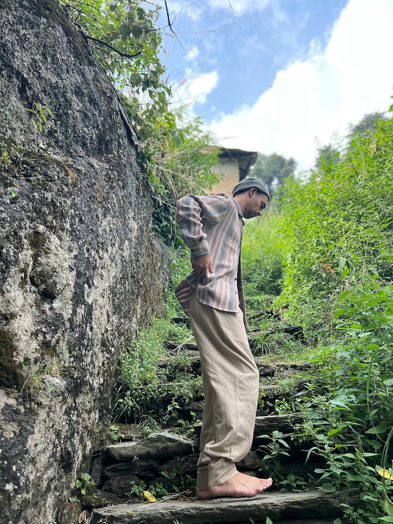 May include: A person wearing a striped shirt and beige pants stands on a set of stone steps in a lush green forest setting.