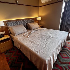 May include: A beige bed with a white linen duvet cover and two pillows. The bed is in a bedroom with a wooden nightstand and a rug with a geometric pattern.