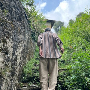 May include: A person wearing a striped shirt and beige pants walks up a stone staircase in a lush green forest setting.