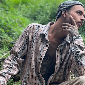 May include: A person wearing a brown and black geometric patterned shirt with a gray beanie. The person is sitting in a grassy area and has a tattoo on their chest.