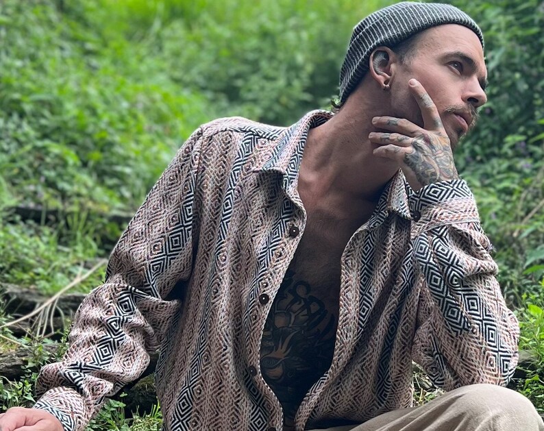 May include: A person wearing a brown and black geometric patterned shirt with a gray beanie. The person is sitting in a grassy area and has a tattoo on their chest.