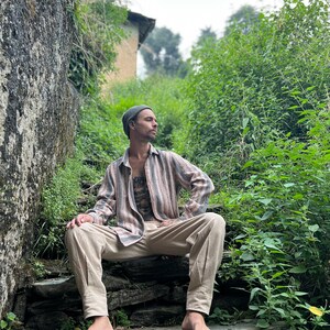 May include: A person wearing a gray beanie and a striped, long-sleeved shirt sits on a stone staircase. The person is wearing beige pants and has a tattoo on their chest. The staircase is surrounded by green foliage.