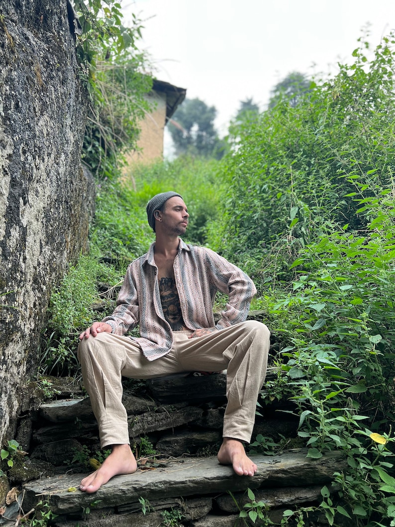 May include: A person wearing a gray beanie and a striped, long-sleeved shirt sits on a stone staircase. The person is wearing beige pants and has a tattoo on their chest. The staircase is surrounded by green foliage.