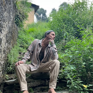 May include: A man wearing a beige shirt with a black and white pattern, beige pants, and a green beanie sits on a stone staircase in a lush green outdoor setting.