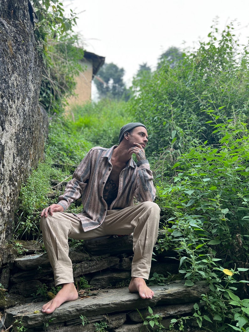 May include: A man wearing a beige shirt with a black and white pattern, beige pants, and a green beanie sits on a stone staircase in a lush green outdoor setting.