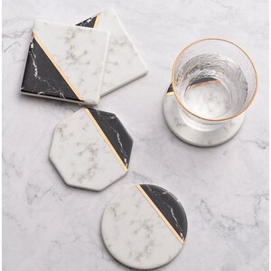 May include: A set of marble coasters in different shapes: square, octagonal, and round. The coasters have a white marble pattern with black sections and a gold stripe. A glass with a gold rim rests on a matching coaster.