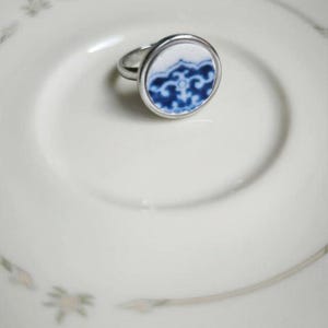 Könnte beinhalten: Ein silberner Ring mit einem runden, weiß-blauen Keramikdesign. Der Ring liegt auf einem weißen Teller mit floralem Muster. Das Keramikdesign zeigt ein blaues, verziertes Muster.