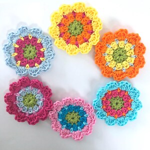 Könnte beinhalten: Eine Sammlung von sieben bunten, gehäkelten Blumenapplikationen. Jede Blume hat einen zentralen Kreis in einer Kontrastfarbe, umgeben von Blütenblättern in Blau-, Rosa-, Orange- und Gelbtönen. Die handgefertigten Blumen sind kreisförmig angeordnet.