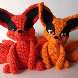 May include: Two handmade crochet plush toys shaped like foxes with multiple tails. One is bright orange and the other is a lighter orange shade. Both feature large pointed ears with black felt accents, black noses, and round eyes.