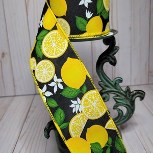 Puede incluir: Un rollo de cinta decorativa con fondo negro adornado con limones amarillos, flores blancas y hojas verdes. La cinta tiene un borde cosido amarillo y se muestra sobre un soporte decorativo.