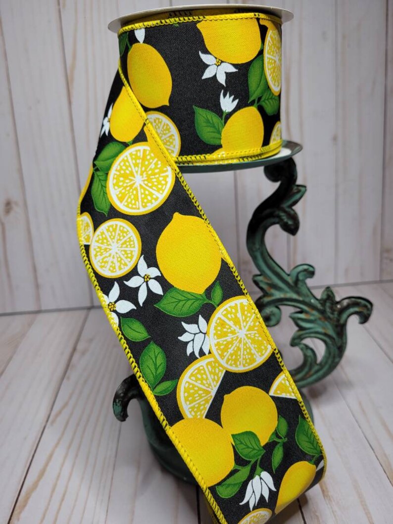 Puede incluir: Un rollo de cinta decorativa con fondo negro adornado con limones amarillos, flores blancas y hojas verdes. La cinta tiene un borde cosido amarillo y se muestra sobre un soporte decorativo.