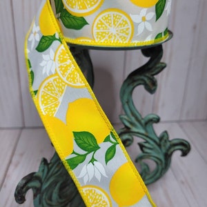 Puede incluir: Un rollo de cinta decorativa con un dise&ntilde;o de lim&oacute;n. La cinta tiene un fondo gris con limones amarillos, flores blancas y hojas verdes. Los bordes est&aacute;n ribeteados en amarillo. La cinta se muestra en un soporte de metal decorativo.
