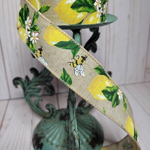 Puede incluir: Un rollo de cinta decorativa con un dise&ntilde;o de lim&oacute;n y abeja. La cinta tiene una textura de arpillera natural y est&aacute; adornada con limones amarillos, hojas verdes, flores blancas y abejas. La cinta se exhibe en un soporte de metal de estilo vintage color turquesa.