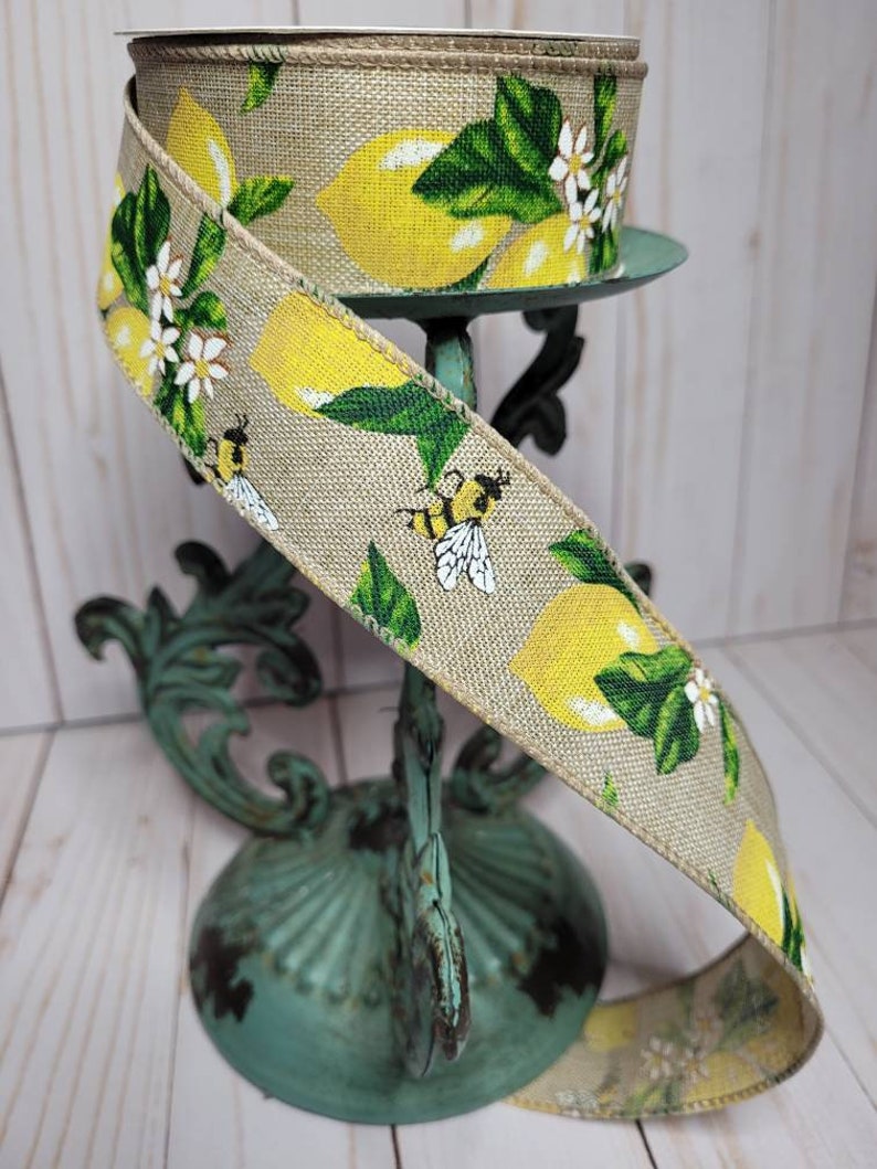 Puede incluir: Un rollo de cinta decorativa con un dise&ntilde;o de lim&oacute;n y abeja. La cinta tiene una textura de arpillera natural y est&aacute; adornada con limones amarillos, hojas verdes, flores blancas y abejas. La cinta se exhibe en un soporte de metal de estilo vintage color turquesa.