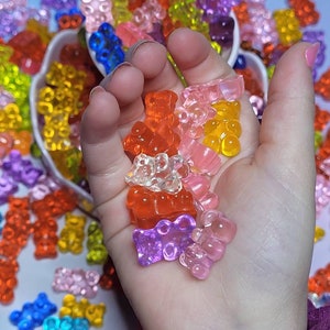 May include: A handful of colorful gummy bear candies in various colors, including red, orange, yellow, pink, purple, and clear. The candies are held in a hand, with more gummy bears visible in the background.