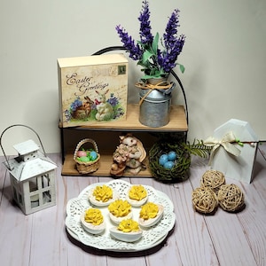 May include: An Easter-themed still life featuring deviled eggs on a white plate, a decorative box with "Easter Greetings" text, and a small metal milk jug with purple flowers. Other decorations include a white lantern, a bunny figurine, and decorative spheres.