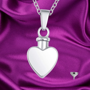 Puede incluir: Un collar con un colgante de cremación en forma de corazón de plata con un pequeño embudo para rellenar. El colgante está en una cadena de plata y se muestra sobre un fondo de satén morado.
