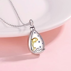 May include: Silver necklace with a teardrop-shaped pendant featuring a gold dolphin jumping over a wave. The pendant is adorned with small blue crystals.