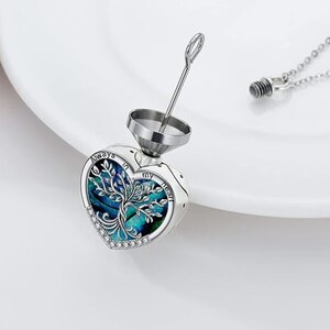 Może przedstawiać: Srebrny naszyjnik z sercowatym wisiorem ozdobionym niebiesko-zielonym drzewem życia. Wisiorek jest grawerowany słowami "Always in my heart". Naszyjnik zawiera mały lejek do napełniania wisiora.
