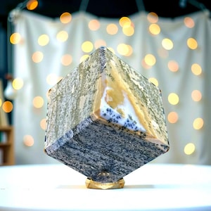 Puede incluir: Una escultura de piedra pulida en forma de cubo con un patrón de color gris, blanco y amarillo. La piedra está montada sobre una base dorada.