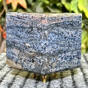 Op de afbeelding: Een blauwe en witte agaatkubus met een glinsterende afwerking, rustend op een gouden metalen standaard met drie poten.