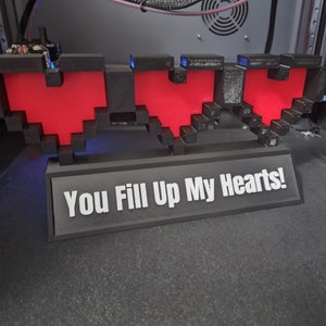 May include: A 3D printed black and red pixelated heart sculpture with a black base that reads "You Fill Up My Hearts!"