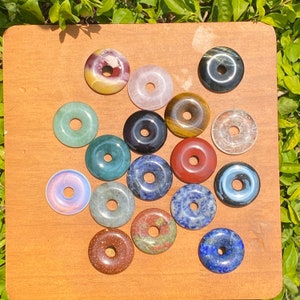 Puede incluir: Surtido de 15 colgantes de piedras preciosas en forma de rosquilla en varios colores, incluidos verde, azul, rojo, rosa, marrón y transparente. Los colgantes están dispuestos sobre una superficie de madera.