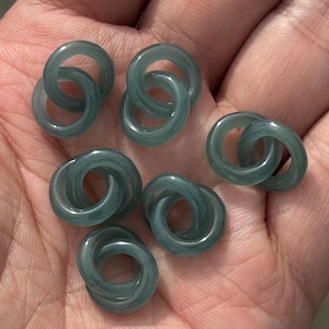May include: A collection of translucent teal jade rings, each featuring two interlocked circles, are shown in a hand. The rings have a smooth, polished finish and a subtle marbled effect. The image suggests jewellery or craft materials.