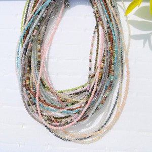 Puede incluir: Una colección de delicados collares de piedras preciosas, cada hebra con un color y tipo de piedra diferente. Los collares están dispuestos en capas, creando una hermosa y colorida exhibición.
