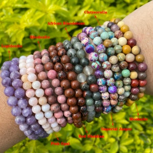 Puede incluir: Una pila de 10 pulseras de cuentas de colores, incluyendo ópalo rosa, obsidiana roja, jaspe imperial, mookaite, crisocola, piedra de sangre africana, rodonita y lepidolita.