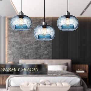 May include: Three blue glass pendant lights with a textured surface hang from black cords in a modern bedroom setting. The lights are illuminated with warm white light bulbs. The text "Warmly Shades" is visible in the image.