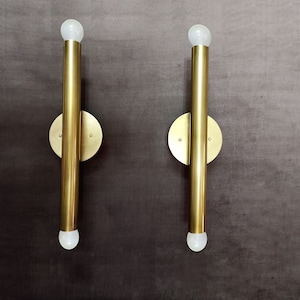 May include: Two gold wall sconces with round, flat backs and a single light bulb at the top of each. The sconces are mounted on a dark gray wall.