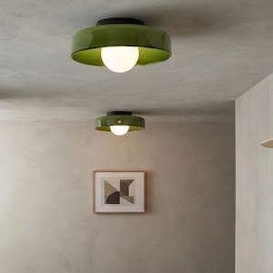 May include: Two olive green flush mount ceiling lights with a circular, glossy shade and a central white spherical glass bulb. The fixtures are mounted on a light-colored ceiling, providing illumination in a hallway with a wooden floor and rug.