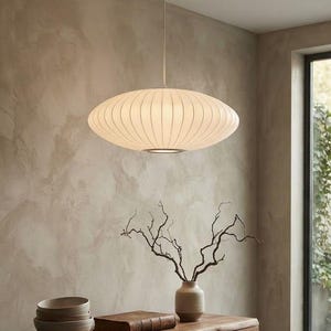 May include: A white, oval-shaped pendant light with a ribbed texture hangs from a thin cord. The light fixture features a rounded, disc-like silhouette that emits a soft, diffused glow, suspended above a wooden surface in a room setting.