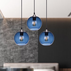 May include: Three blue glass globe pendant lights hang from black cords in a modern bedroom setting. The lights are suspended over a white bed with a white headboard and a white bench at the foot of the bed. The text "Warmly Shades" is visible in the image.