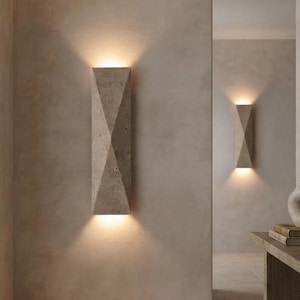 May include: A rectangular wall sconce with a textured, stone-like finish and a geometric, faceted design. The fixture emits a warm, upward and downward glow, casting soft light against the wall surface to create a distinct, vertical illumination.