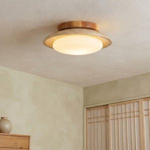 May include: A flush mount ceiling light fixture featuring a circular wooden base with a natural grain finish, topped by a wide, shallow, textured beige shade and a central, glowing, frosted white glass dome that emits a soft, warm light.