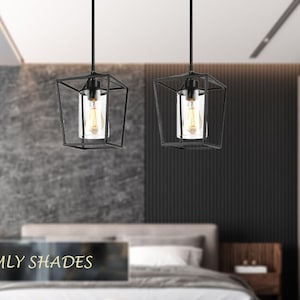 May include: Two black caged pendant lights with clear glass shades hang over a bed in a modern bedroom. The lights are suspended from the ceiling and have a simple, industrial design.