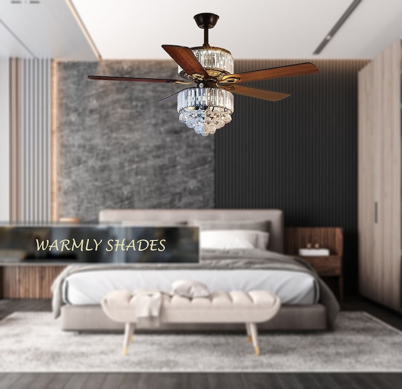 Puede incluir: Un ventilador de techo con aspas de madera y una l&aacute;mpara de ara&ntilde;a de cristal. El ventilador est&aacute; en un dormitorio con una cama y un banco.